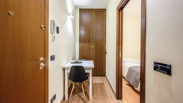 A narrow hallway with a wooden door, a white table with a black chair, a bedside table with a lamp, and a bed with a gray blanket. - Emilia Studios 2 (Modena)