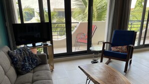Una sala de estar con un sofá, una mesa de centro de madera, un sillón azul, un televisor sobre un mueble bajo y un balcón con vistas a palmeras y edificios.