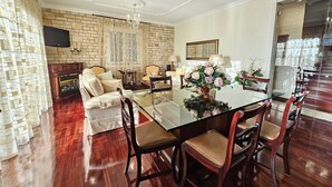 A spacious living room with a large glass dining table surrounded by wooden chairs, a fireplace, and a television mounted on the brick wall.