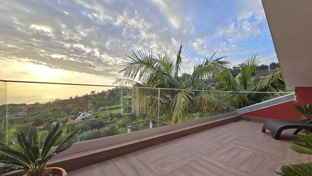 A balcony with a wooden floor, a glass railing, and a view of a green landscape with houses.