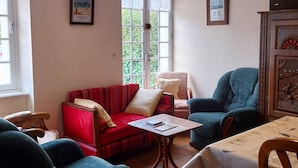 Una sala de estar acogedora con un sofá rojo, un sillón azul y una mesa de madera. Hay dos cuadros enmarcados en la pared, una ventana con cortinas de encaje y un ventilador de techo.