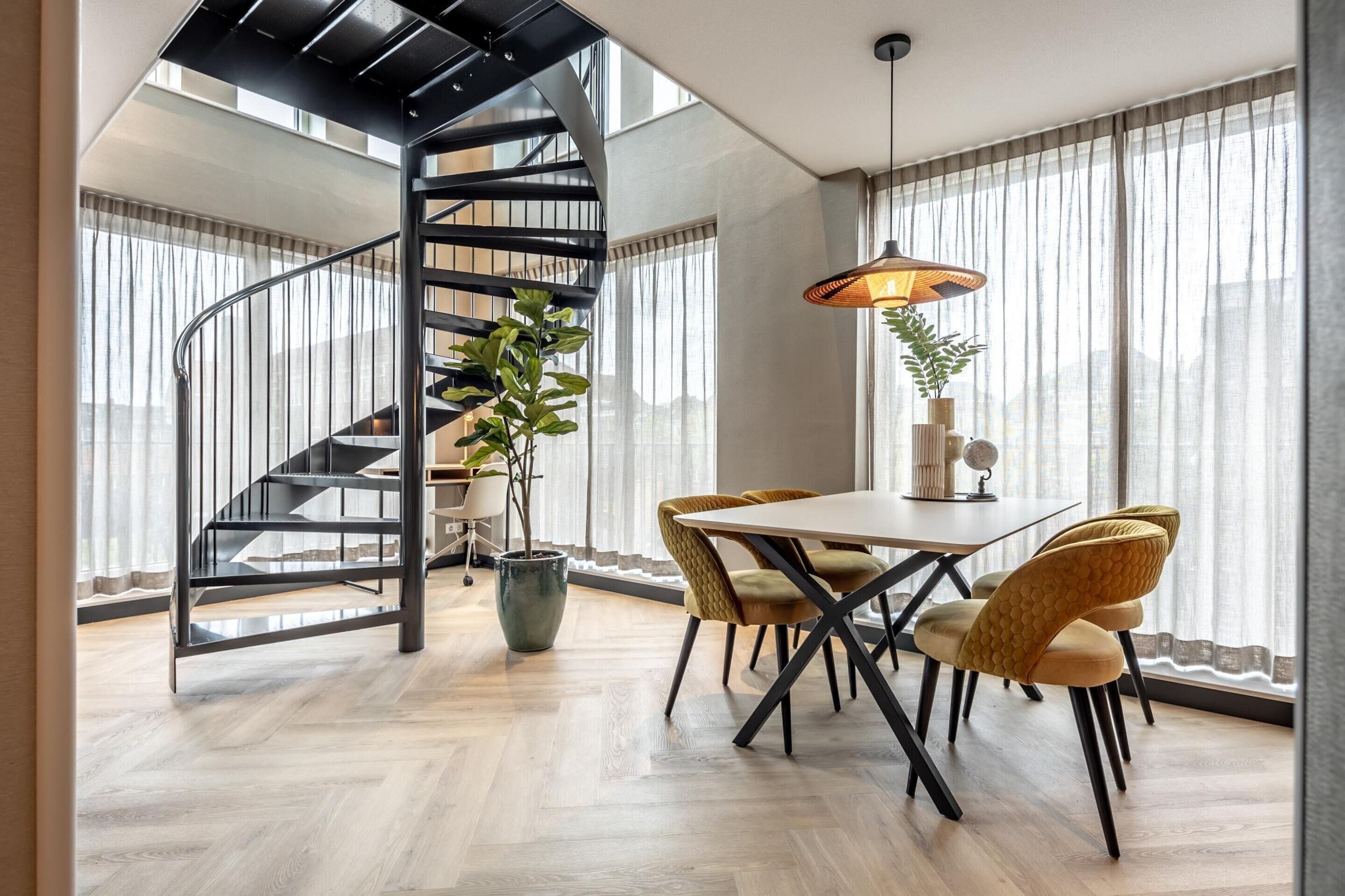 Un interior moderno con una escalera espiral, un comedor con mesa y sillas, y ventanas grandes con cortinas translúcidas.