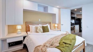 Une chambre d’hôtel moderne avec un grand lit, des tables de chevet avec des lampes, une tête de lit en bois et, au fond, une petite cuisine.