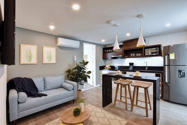 Una cocina moderna con una isla central, taburetes de bar y un sofá gris en una sala de estar.