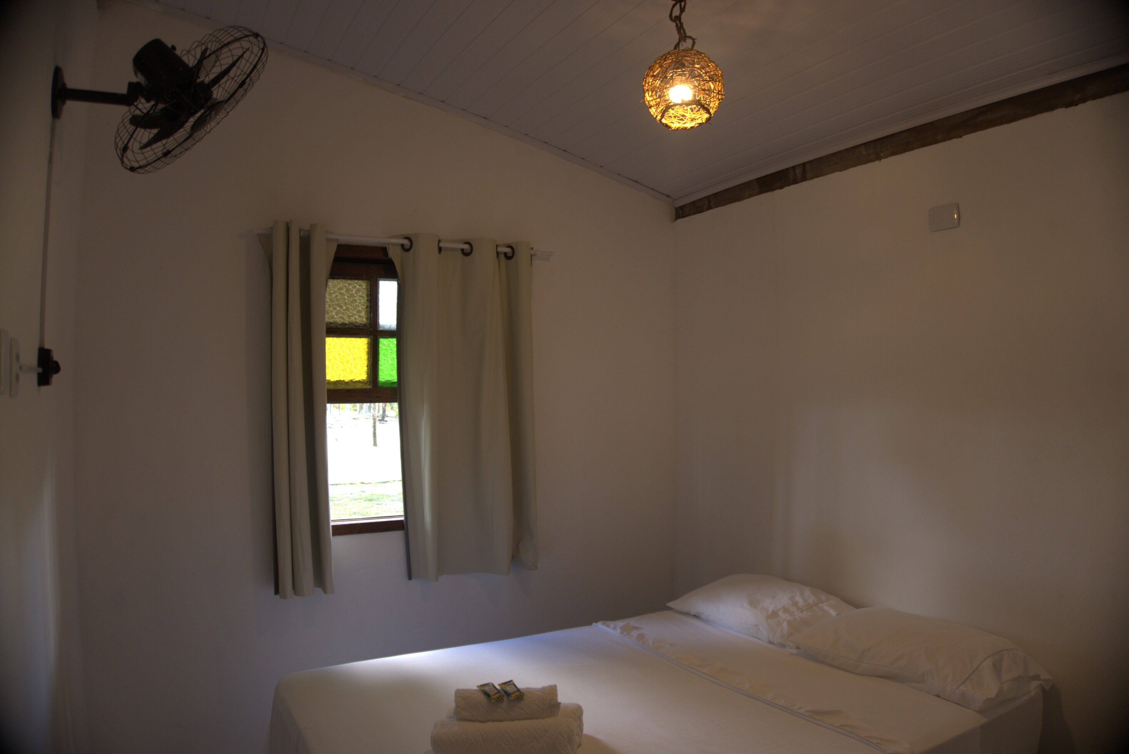 Quarto com cama, janela com cortinas, ventilador de teto e luminária pendente.