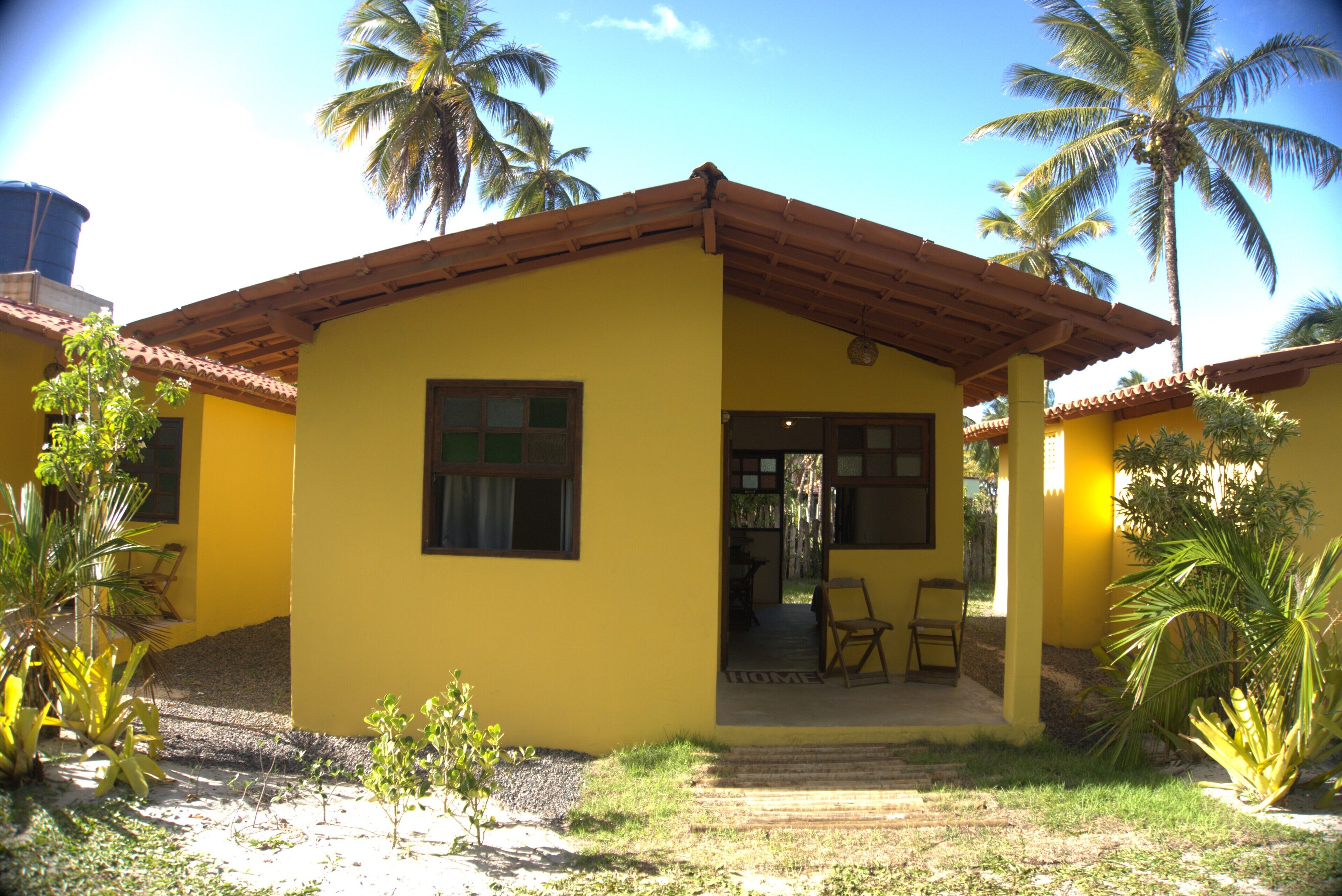 A yellow building with a tiled roof, a covered porch, and palm trees in the background.