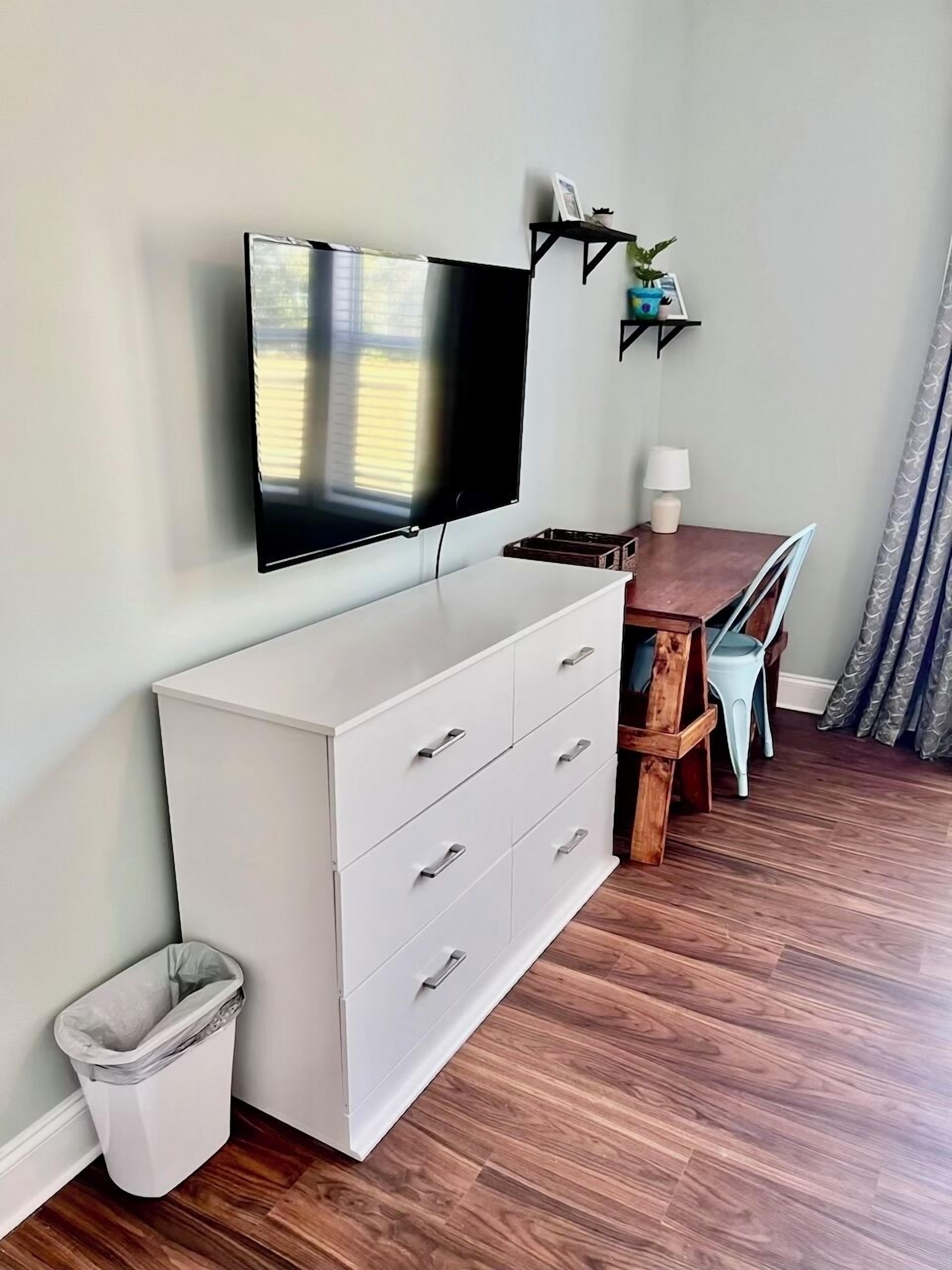 A room with a flat-screen TV mounted on the wall, a white dresser with drawers, a small table with a lamp, and a trash bin.