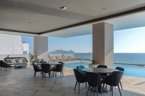 Relax by the pool in a stylish outdoor lounge with a sofa, chairs, and a table, offering breathtaking views of the sea and city.