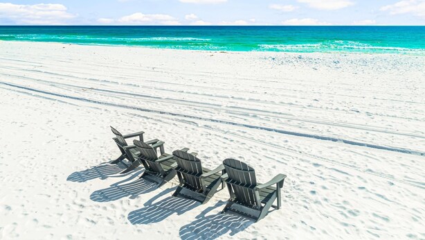 Ein Strand mit weißem Sand und türkisblauem Wasser. Vier Adirondack-Stühle sind am Strand aufgereiht.