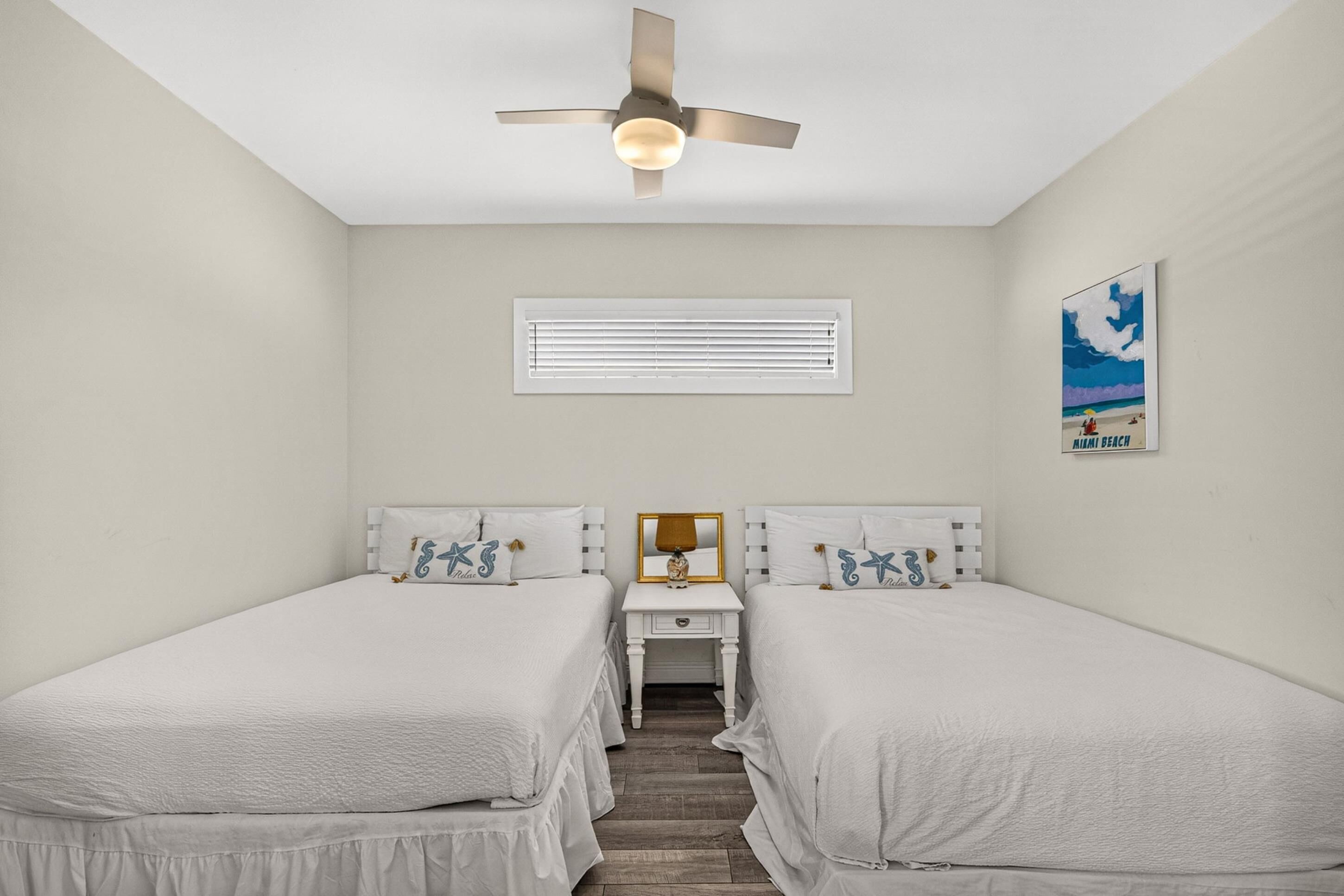 Un dormitorio con dos camas, un ventilador de techo, una mesita de noche, un espejo, una ventana con persianas y un cartel de Miami Beach.