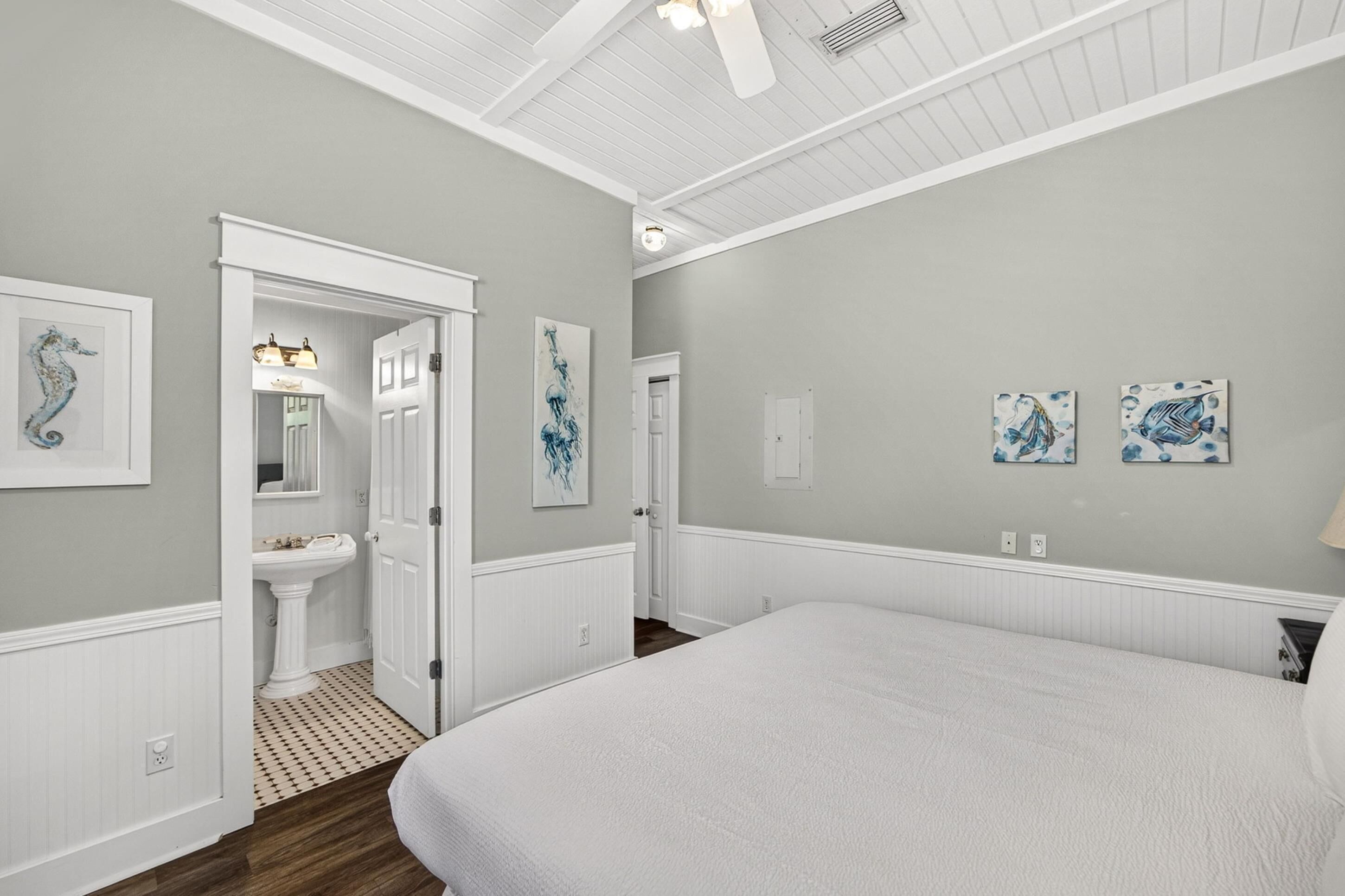 A bedroom with a large bed, a white vanity, a door leading to another room, and wall-mounted artwork.