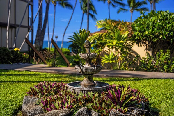 A stone fountain with a tiered design, surrounded by a well-maintained garden with various plants and rocks.