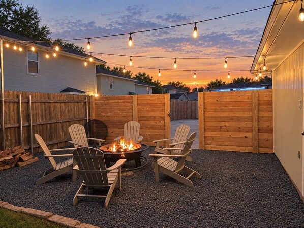 A backyard with a fire pit and Adirondack chairs, surrounded by a wooden fence and string lights. - Large Group Retreat: Two Homes, Sleeps 16 (College Station)