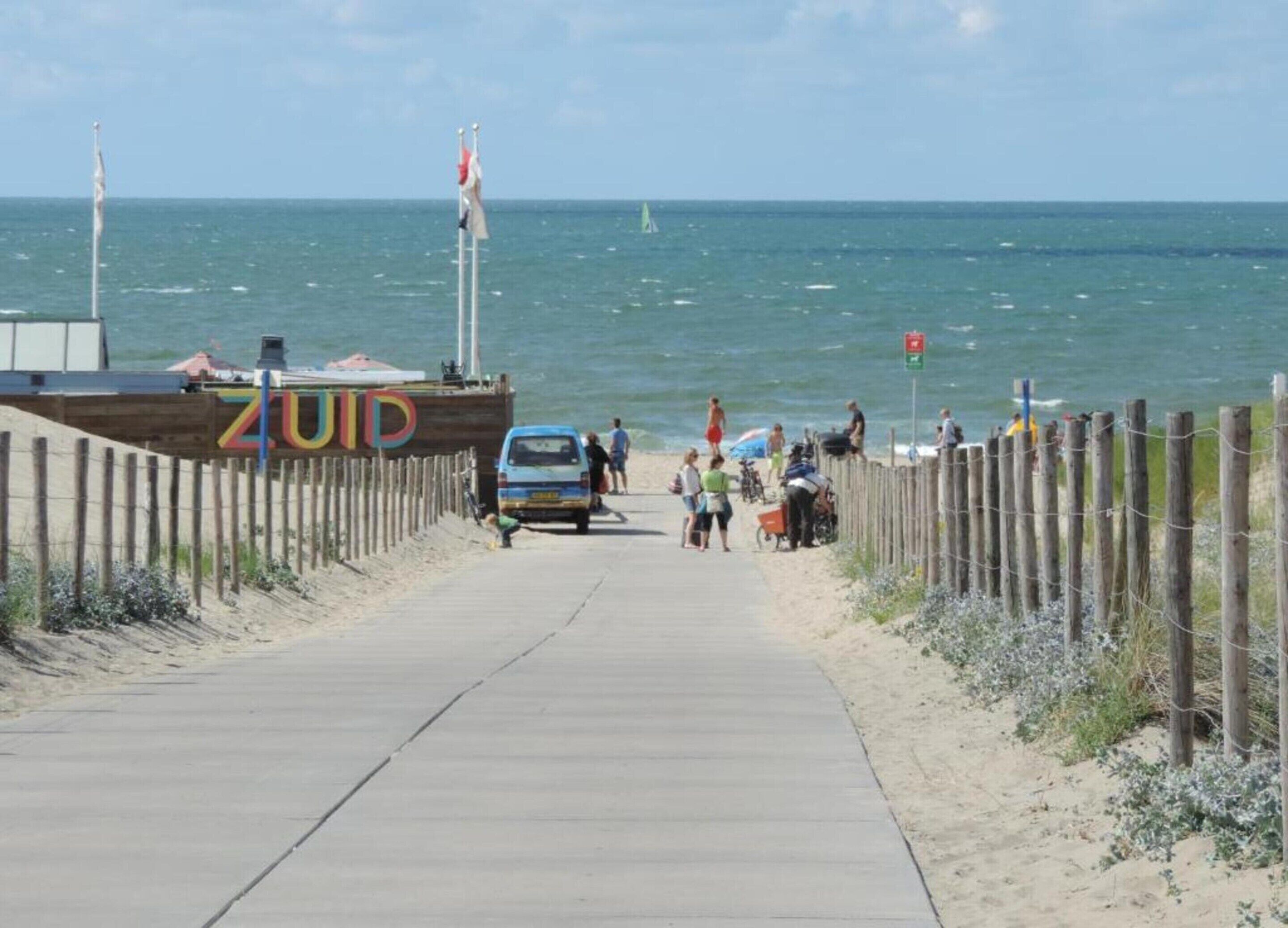 A beachside walkway with a wooden fence, a sign reading "ZUID", and a blue vehicle parked on the path.