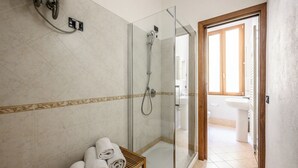 A modern bathroom with a glass shower enclosure, a wooden shelf with towels, and a view into the adjacent bathroom. - Emilia Studios 3 (Modena)
