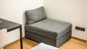 A small, gray sofa in a corner of a room with a wooden floor and a white wall. - Emilia Studios 3 (Modena)