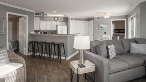 Un salon moderne avec un canapé gris, une petite table avec une lampe et un coin cuisine doté de meubles blancs et d’un tabouret de bar.