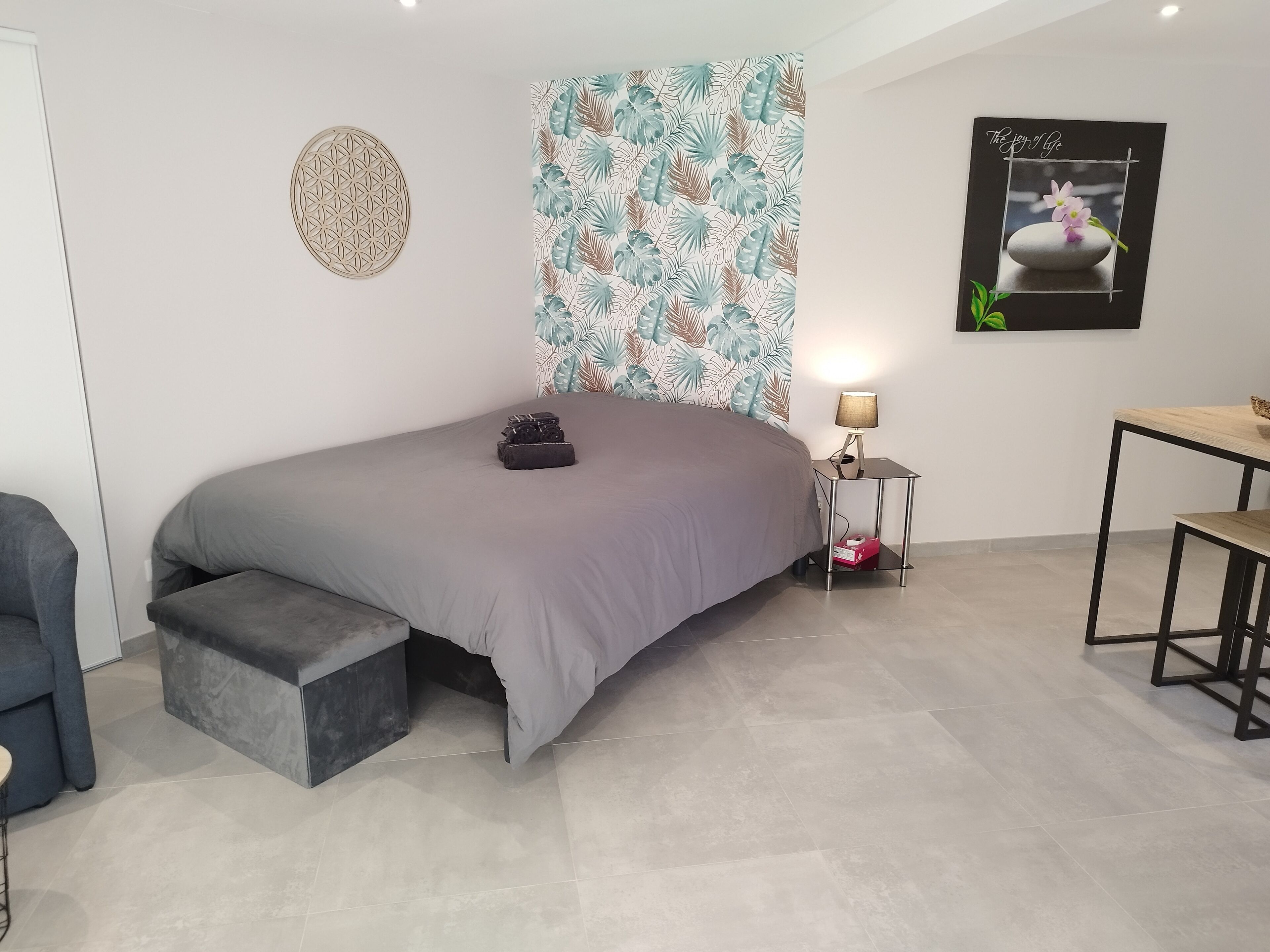 A modern bedroom with a bed, a side table with a lamp, a small desk, and a wall with a tropical leaf pattern.