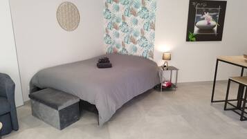 A modern bedroom with a bed, a side table with a lamp, a small desk, and a wall with a tropical leaf pattern.