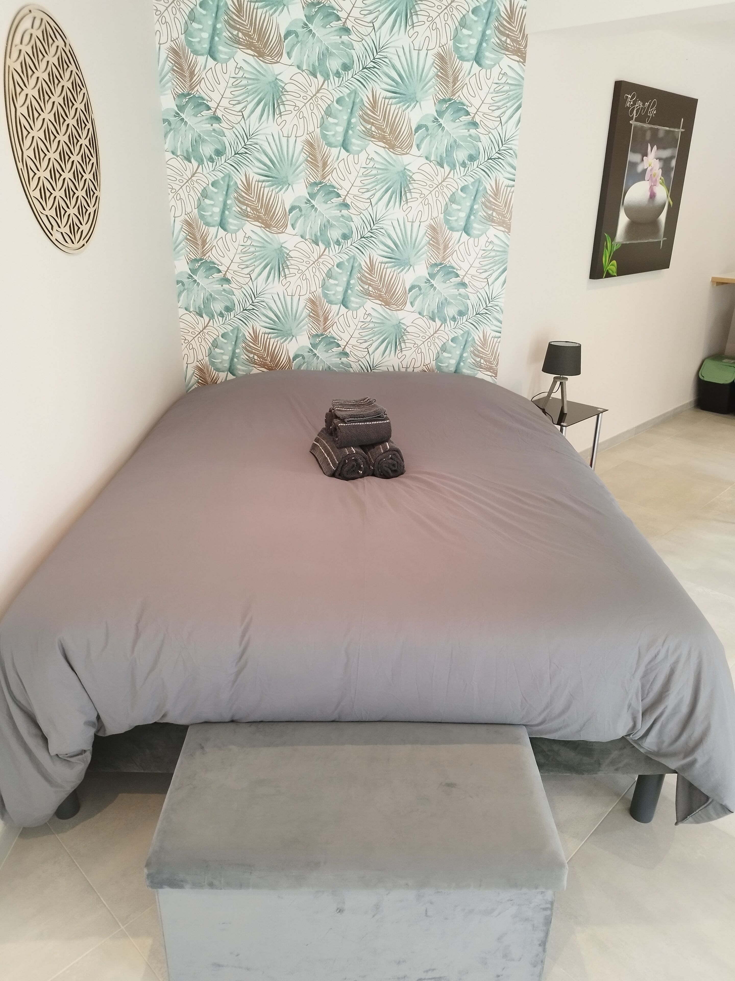 Un lit bien fait, recouvert d’une couverture grise, avec une serviette pliée et un banc au pied du lit.