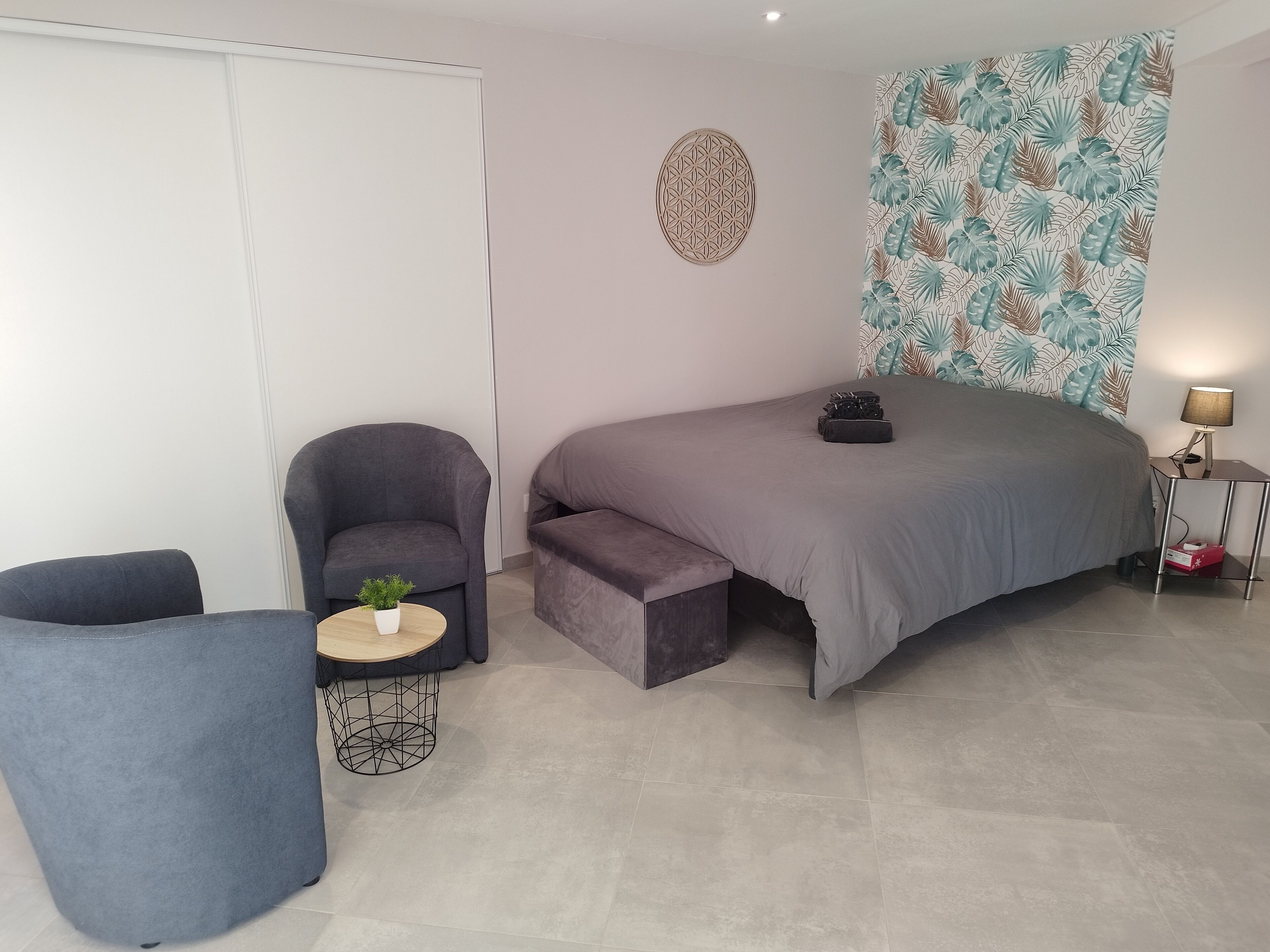Een moderne slaapkamer met een bed, een stoel, een klein tafeltje met een plant en een muur met een tropisch bladmotief.