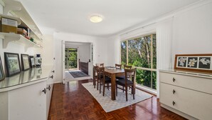 A dining area with a wooden table and chairs, a white cabinet with drawers, and a large window with a view of greenery.