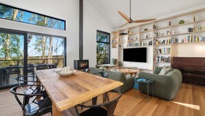Una sala de estar moderna con una mesa de comedor de madera, un sofá verde y una chimenea. Amplias ventanas ofrecen vistas a árboles y un balcón.