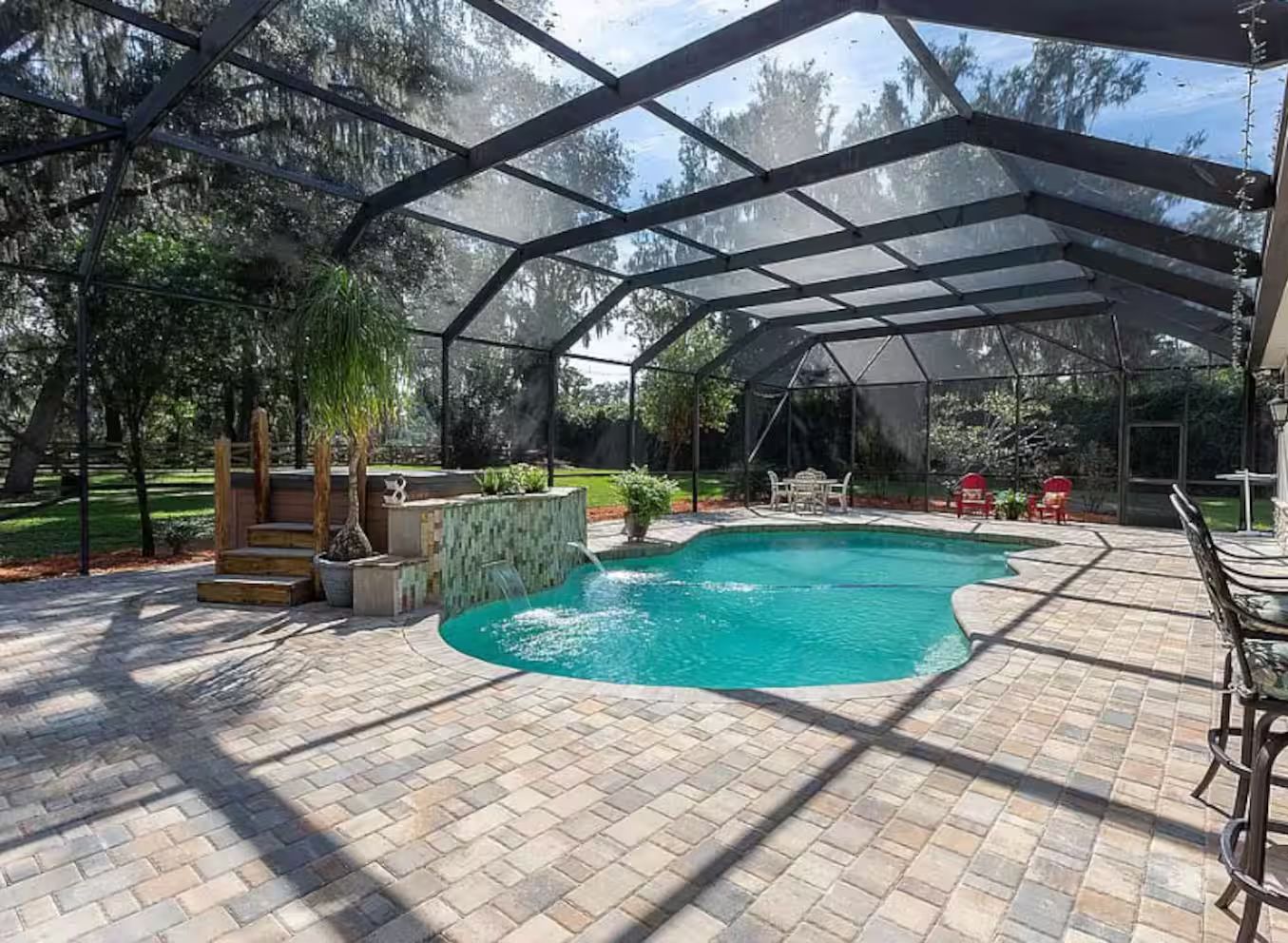 Une piscine extérieure couverte, agrémentée d’une cascade, entourée d’une terrasse en briques et d’un espace aménagé avec des meubles de jardin.