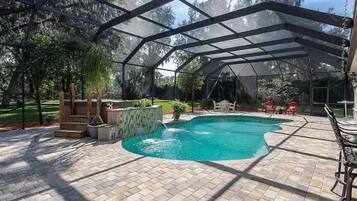 A covered outdoor swimming pool with a waterfall feature, surrounded by a brick patio and a screened area with outdoor furniture.