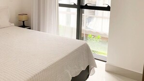 A bedroom with a bed, a nightstand, a window with a view of greenery, and a door with a handle. - Fortune by Day by Day (Joao Pessoa)