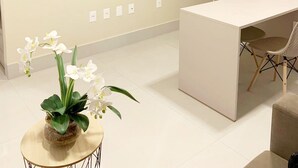 A modern, minimalist room with a flat-screen TV mounted on the wall, a small round table with a potted orchid, a white desk with two chairs, and a door in the background. - Fortune by Day by Day (Joao Pessoa)