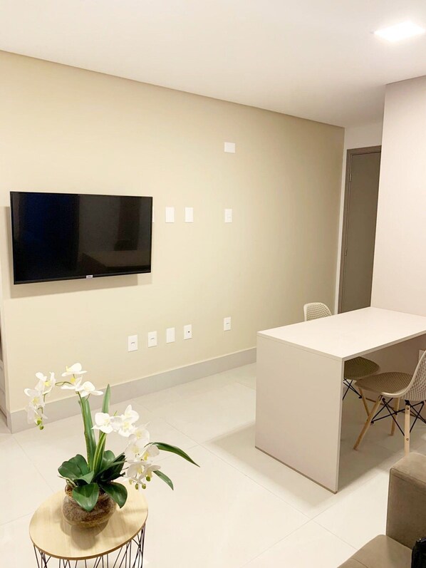 A modern, minimalist room with a flat-screen TV mounted on the wall, a small round table with a potted orchid, a white desk with two chairs, and a door in the background. - Fortune by Day by Day (Joao Pessoa)