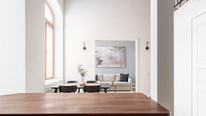 A modern kitchen with a wooden countertop and dark cabinetry, a dining area with a table and chairs, and a living room with a sofa and a large abstract painting.