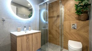 Ein modernes Badezimmer mit Holzpaneelen, einer Duschkabine aus Glas, einer weißen Toilette und einem runden Spiegel mit beleuchtetem Rand.
