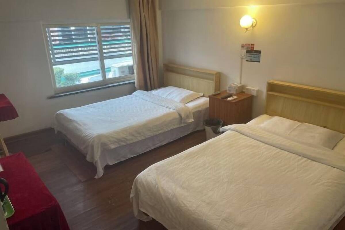 A hotel room with two beds, a window with blinds, a bedside table with a lamp, and a red chair.