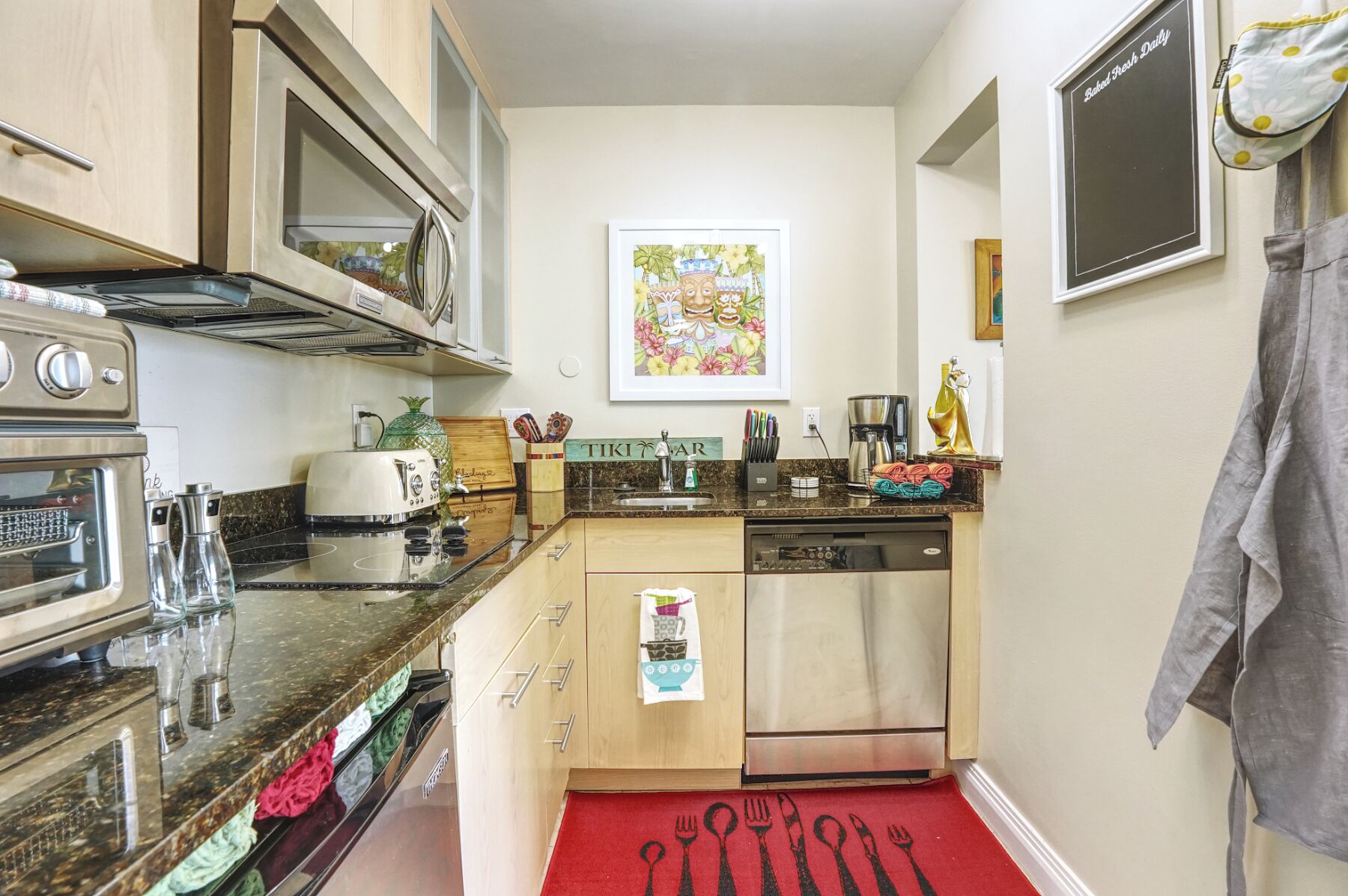 A compact kitchen with a toaster, a coffee maker, and a dishwasher. There is a wooden cabinet above the sink and a framed artwork on the wall.