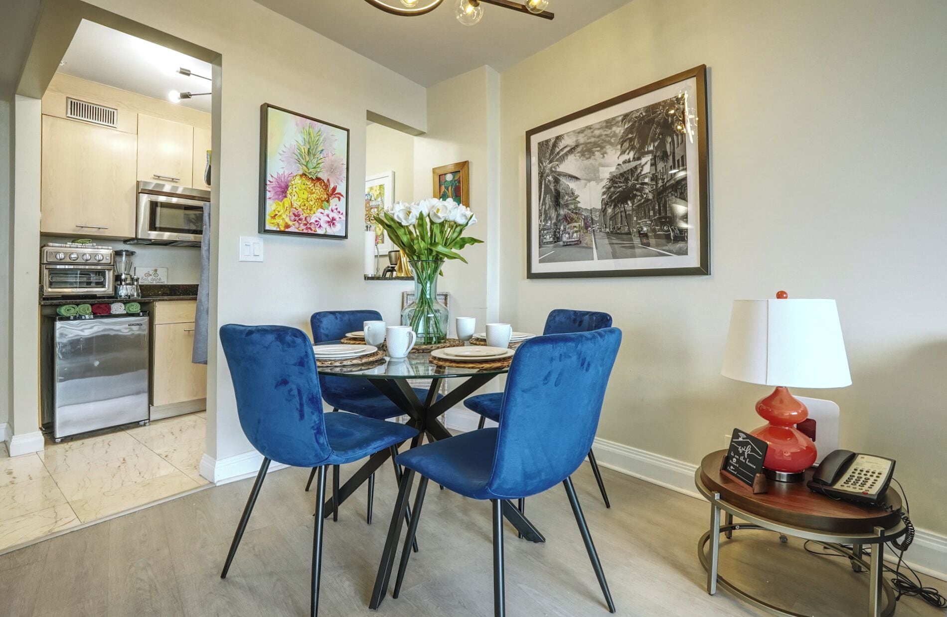 Un comedor con una mesa de vidrio, sillas azules, un florero con flores y una lámpara roja.