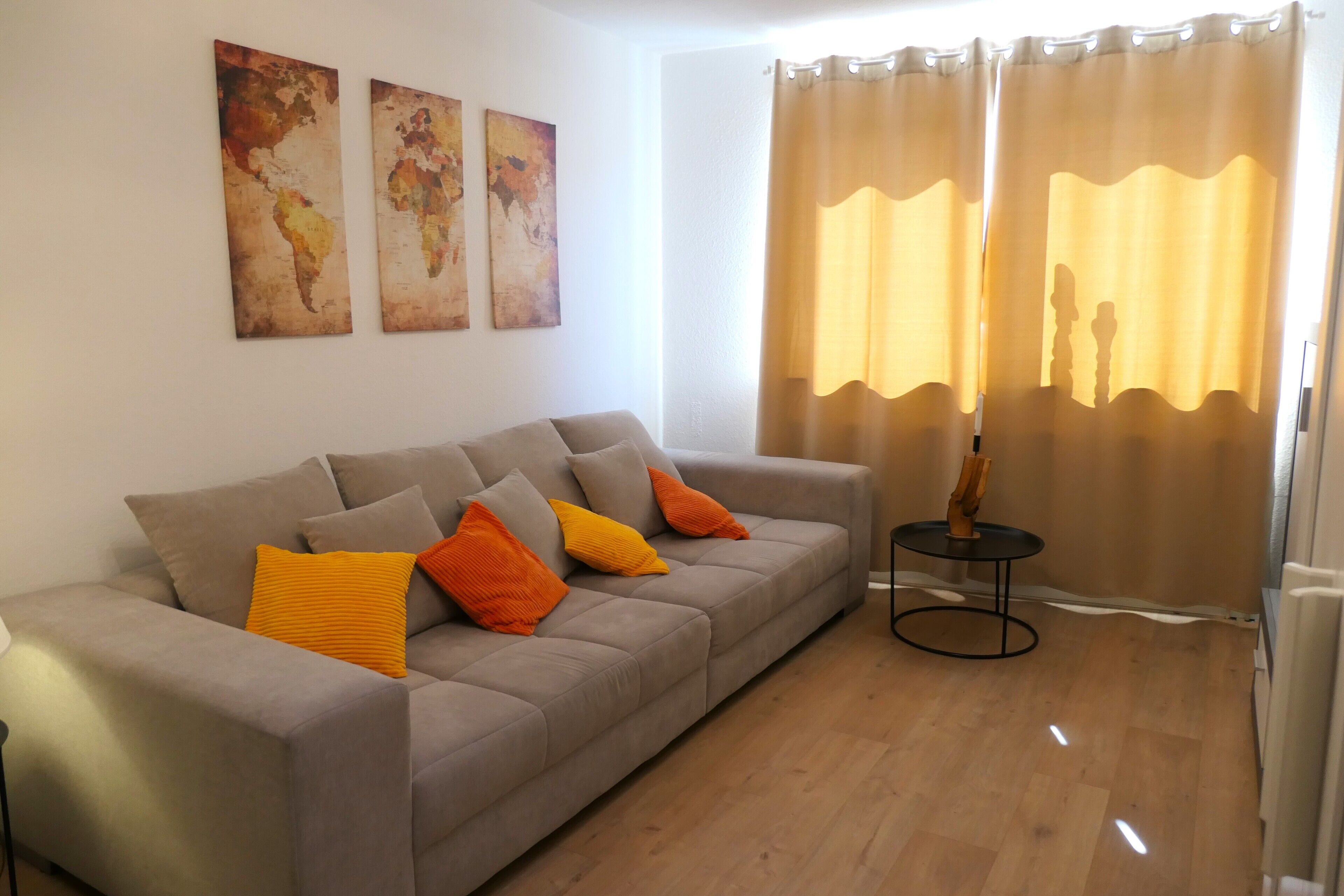 Ein Wohnzimmer mit grauem Sofa, orangefarbenen Kissen, einem kleinen runden Tisch und einer Stehlampe. An der Wand befinden sich abstrakte Gemälde.
