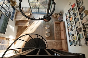 Una scala a chiocciola conduce a una camera da letto soppalcata con una grande finestra, una libreria e un angolo salotto accogliente.