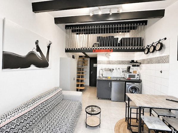 Une cuisine compacte équipée d’un lave-linge, un coin repas avec une table et des bancs, un canapé à motifs et un escalier menant à un niveau supérieur.