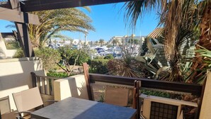 Un patio exterior con mesa y sillas, rodeado de palmeras y vistas a un puerto deportivo con barcos.