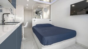 Una habitación de hotel moderna con una cama, una kitchenette pequeña, un espejo y un televisor montado.