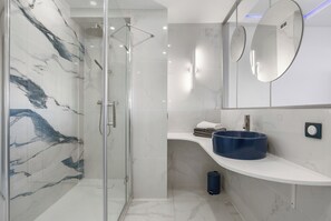 A modern bathroom with a glass-enclosed shower, a marble wall, a blue sink, and a mirror.