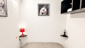Een slaapkamer met een bed, een rode lamp, een lijst met een afbeelding en een plankje met een decoratief object.