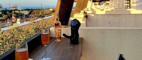 A wooden deck with two glasses of amber liquid, a bottle, and a black cat statue.