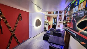 A modern hotel room with a red accent wall, a circular light fixture, a bed with a geometric-patterned headboard, and a wall-mounted TV displaying sports content.