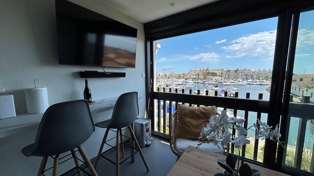 A modern living room with a TV, two chairs, a wooden table, and a balcony with a view of a marina and buildings. - Naturiste Cap d'Agde Port Vénus 212 (Agde)