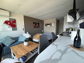 Una sala de estar moderna con un sofá, una mesa de centro de madera, una zona de comedor con sillas, una cocina con microondas y un espejo grande en la pared.