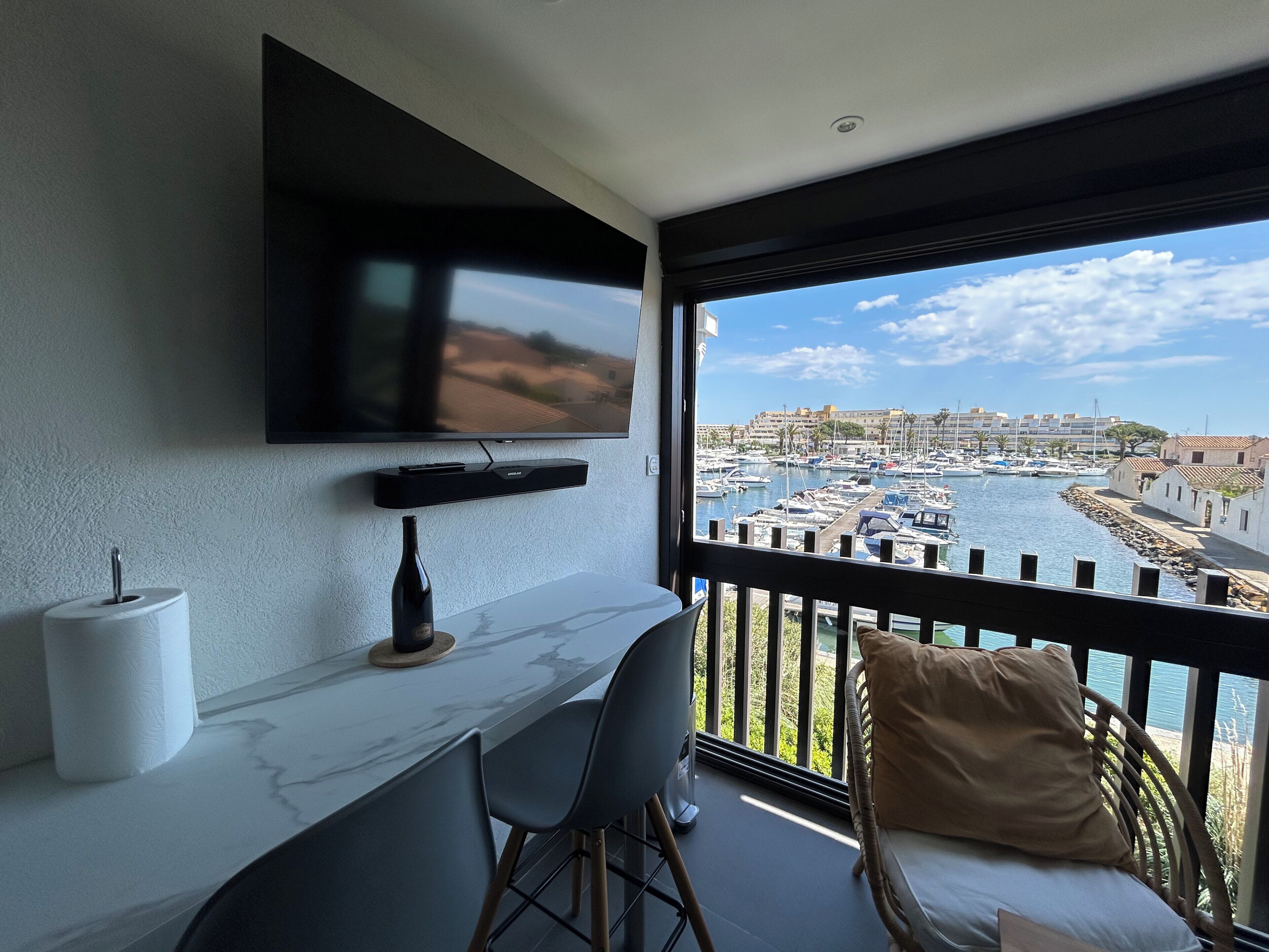A balcony with a TV, a marble table, and chairs, overlooking a marina with boats and buildings.