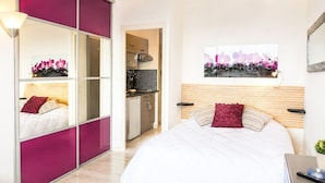 Een slaapkamer met een bed, nachtkastje, spiegel en uitzicht op de keuken.